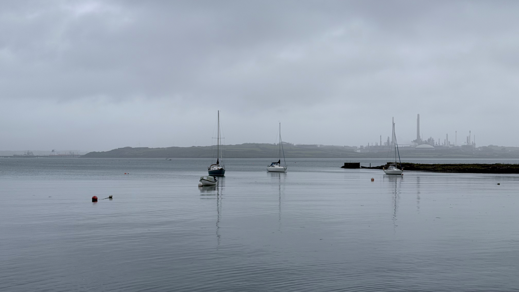 A calm waterscape features several sailboats, distant industrial structures, and a cloudy sky.