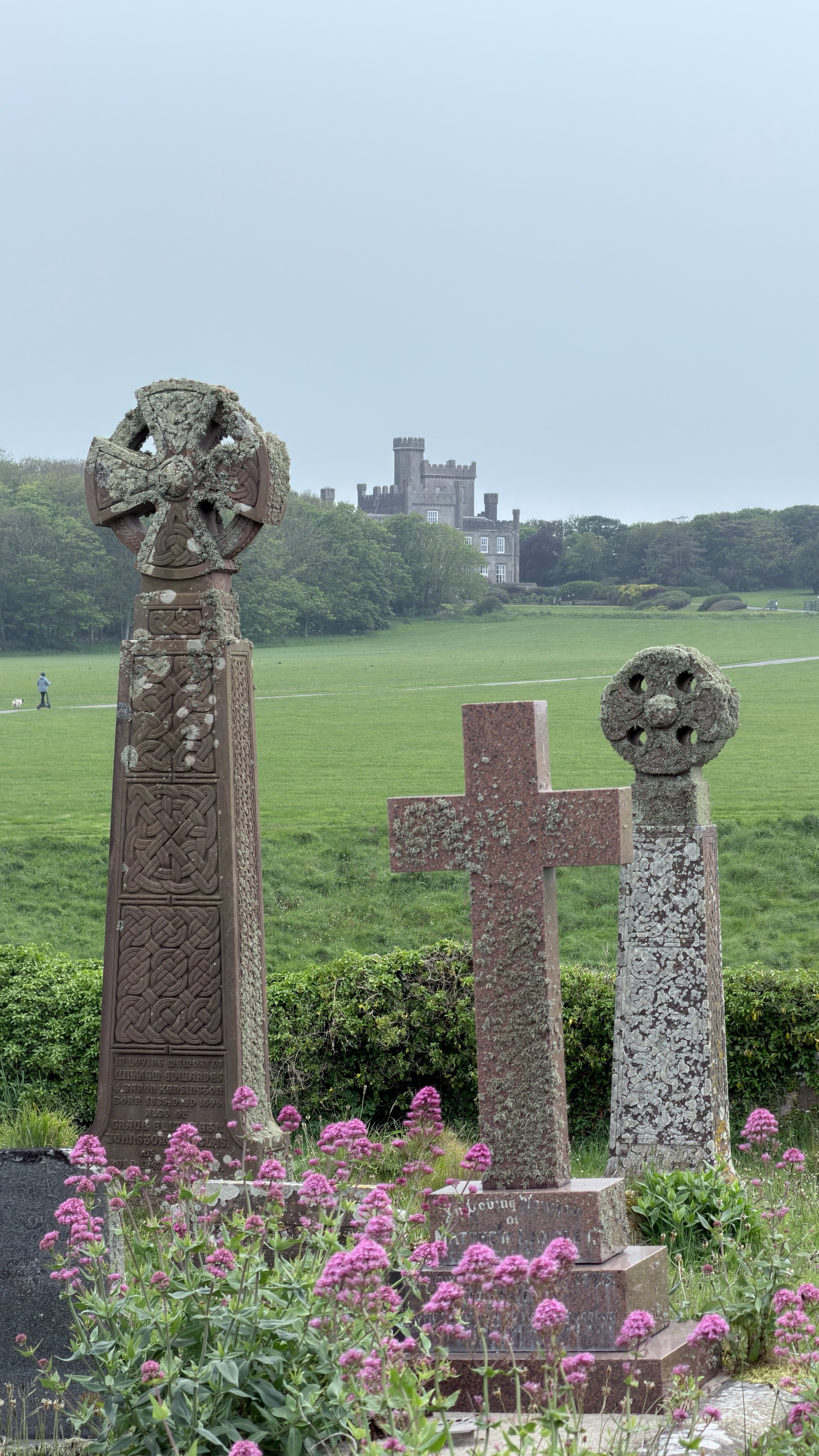 Three intricately carved standing stones with crosses are set against a grassy landscape with a distant view of a castle.
