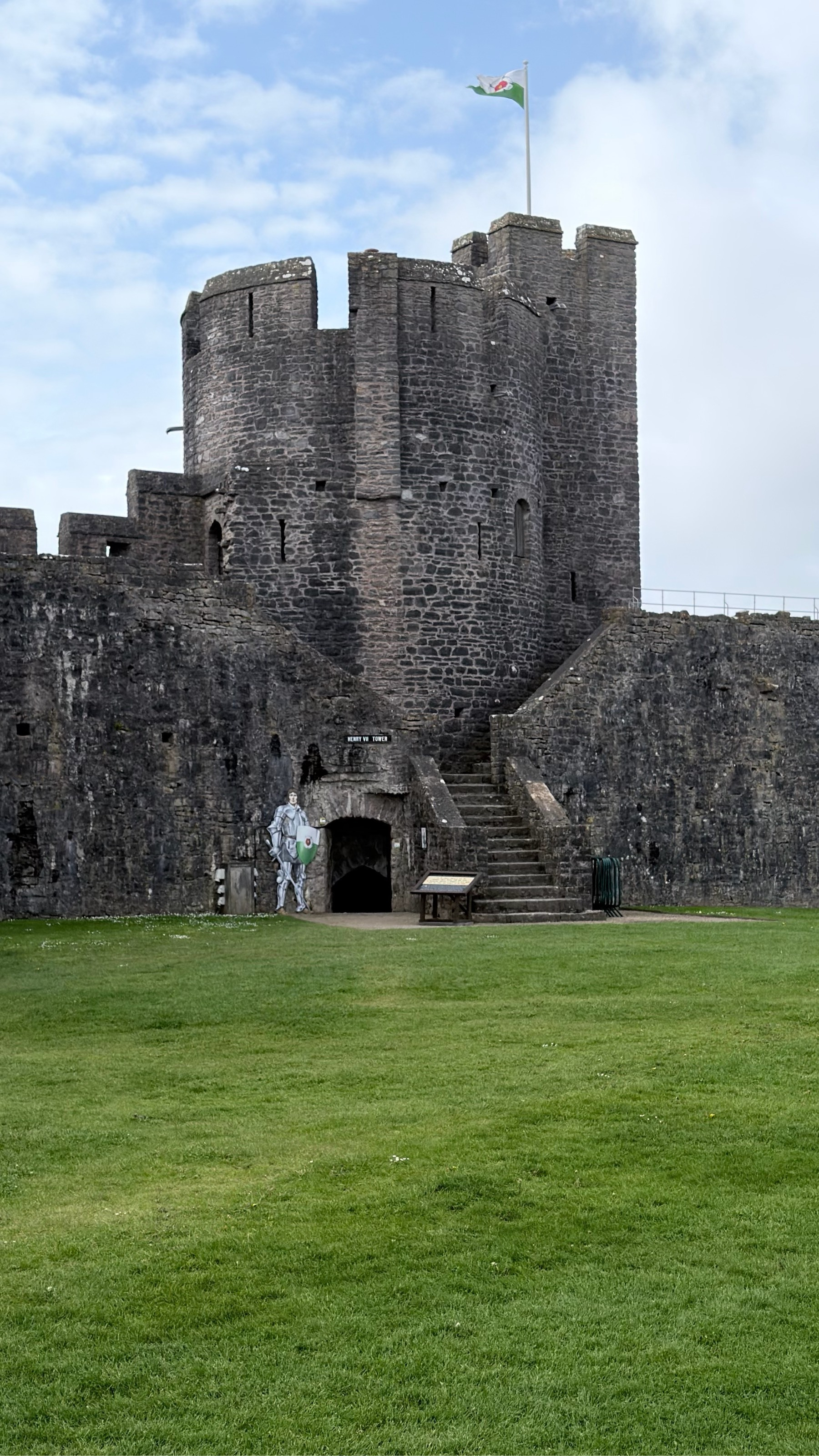 A stone castle with a tower and battlements stands against a cloudy sky, featuring a flag at the top and an arched entrance near a grassy area.