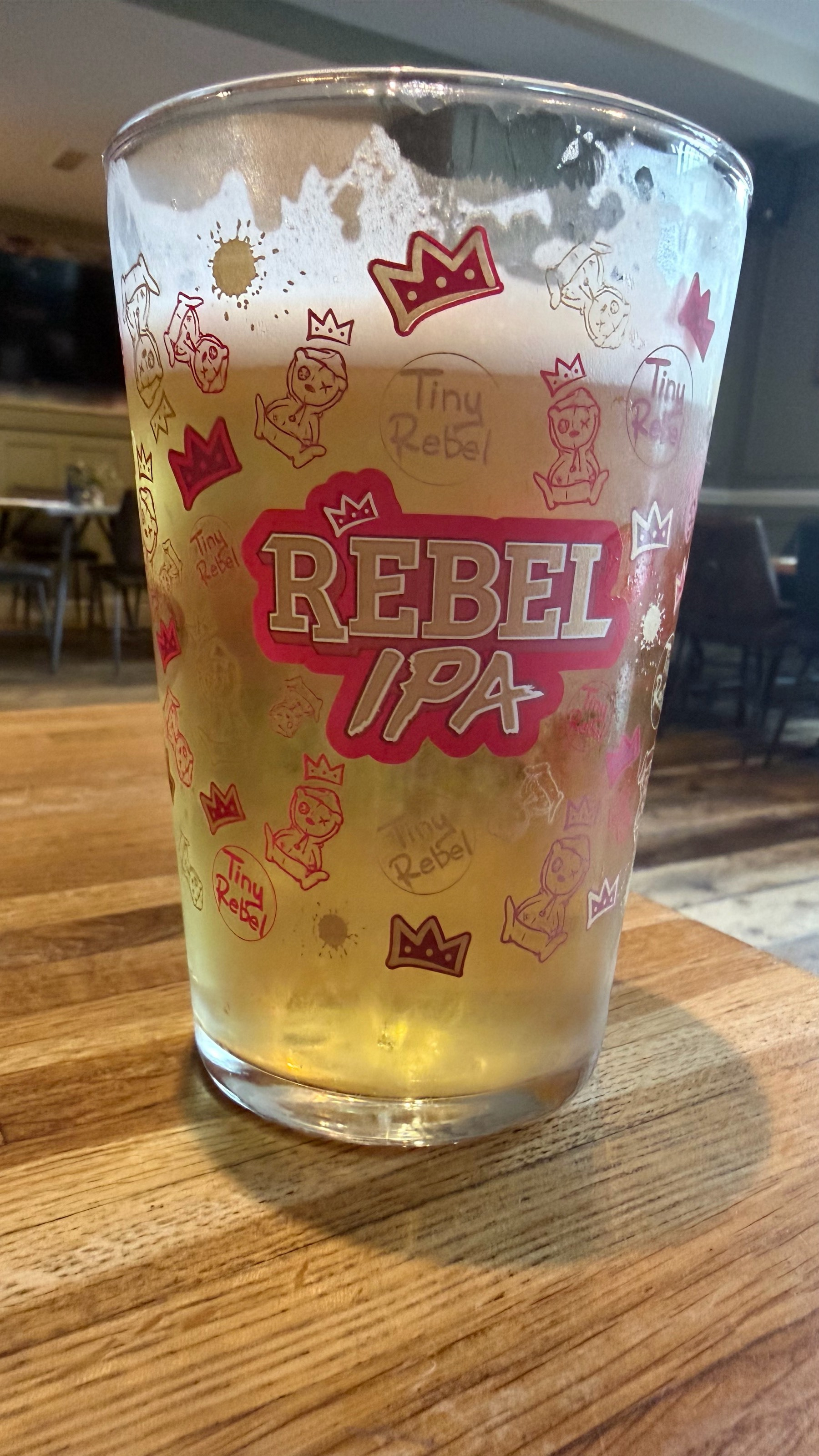 A pint glass of light beer with Rebel IPA and playful graphics is resting on a wooden table.
