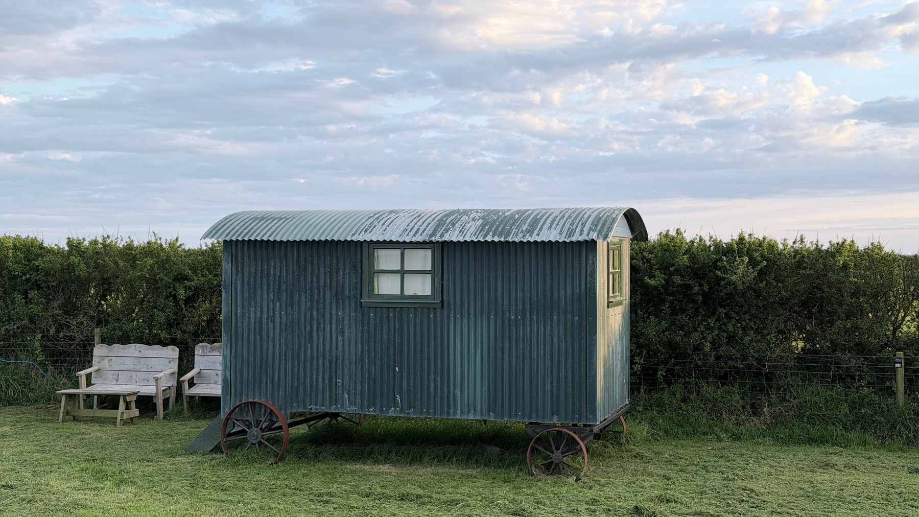 A small, rustic wheeled hut stands on a grassy field with two benches nearby and a hedge in the background.