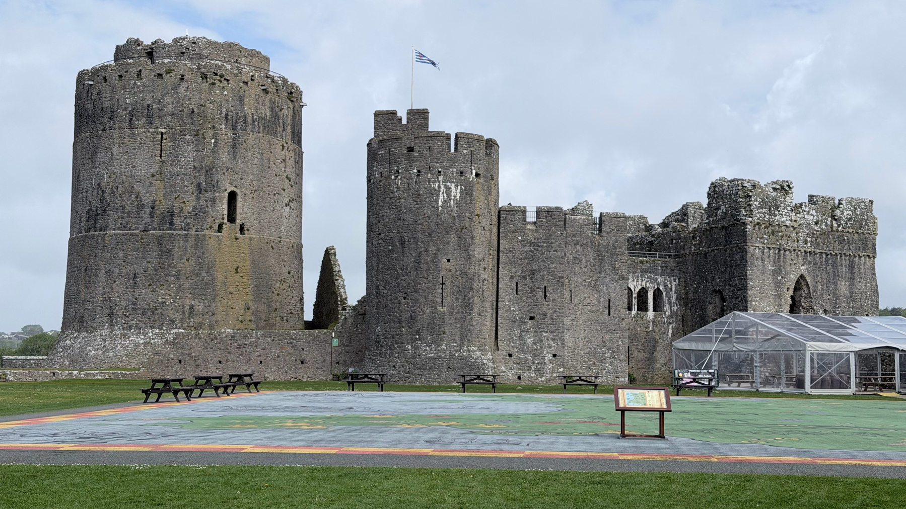 A large medieval castle with round towers and battlements sits on a lush green lawn under a cloudy sky.