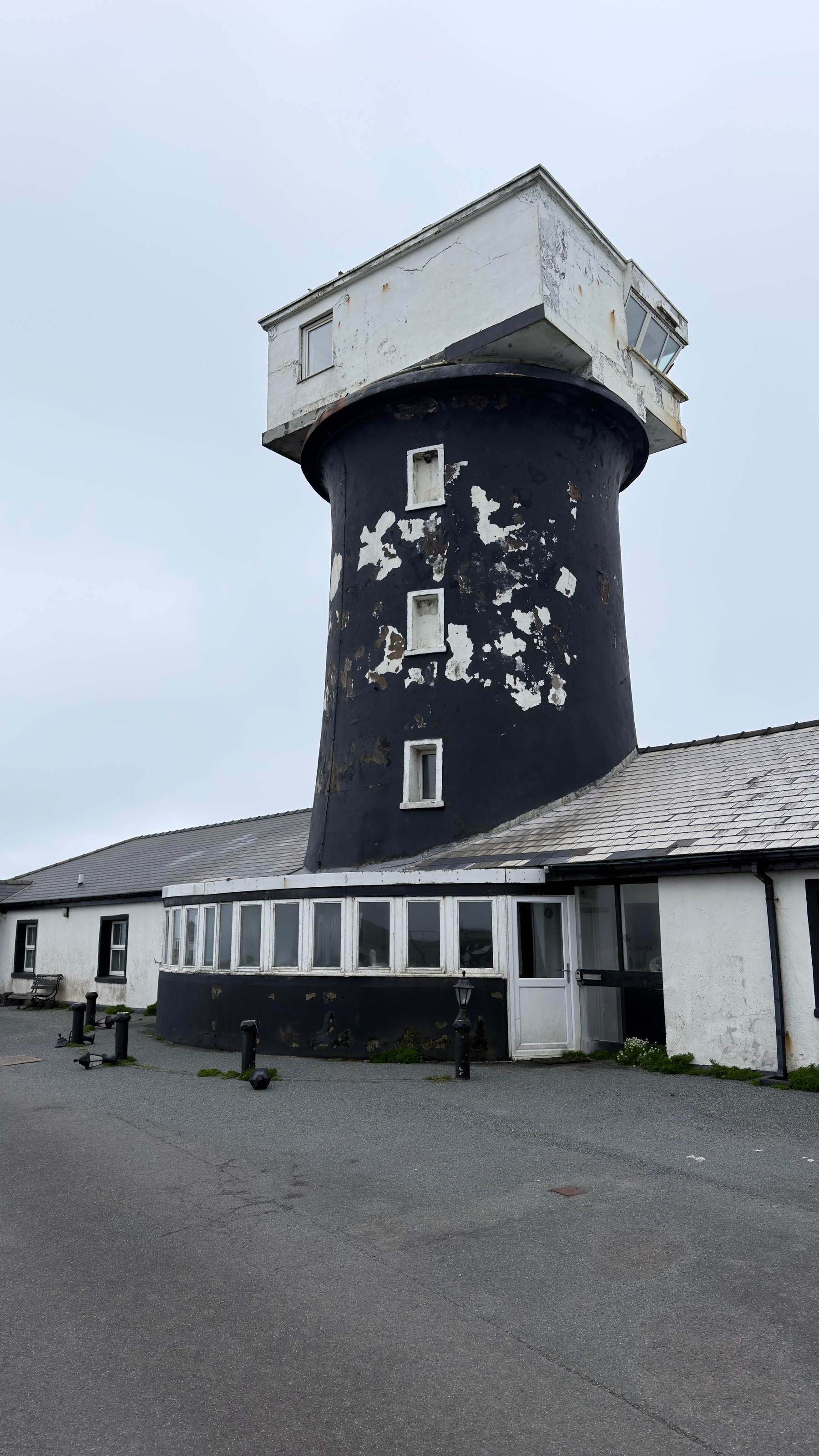A tall, weathered tower with a small, white structure on top is attached to a low, sprawling building on a cloudy day.