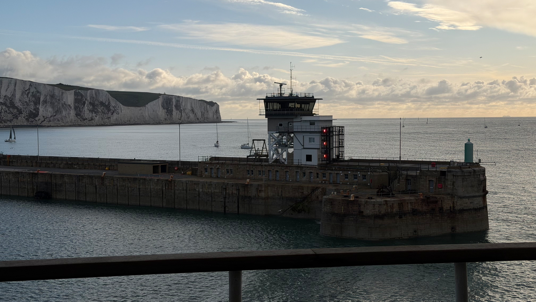 A maritime control tower is situated on a pier with boats sailing nearby and white cliffs in the background.