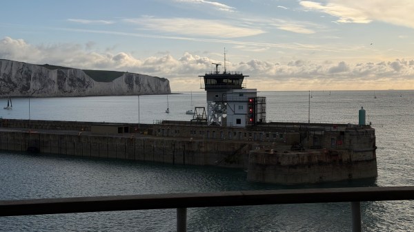 A maritime control tower is situated on a pier with boats sailing nearby and white cliffs in the background.
