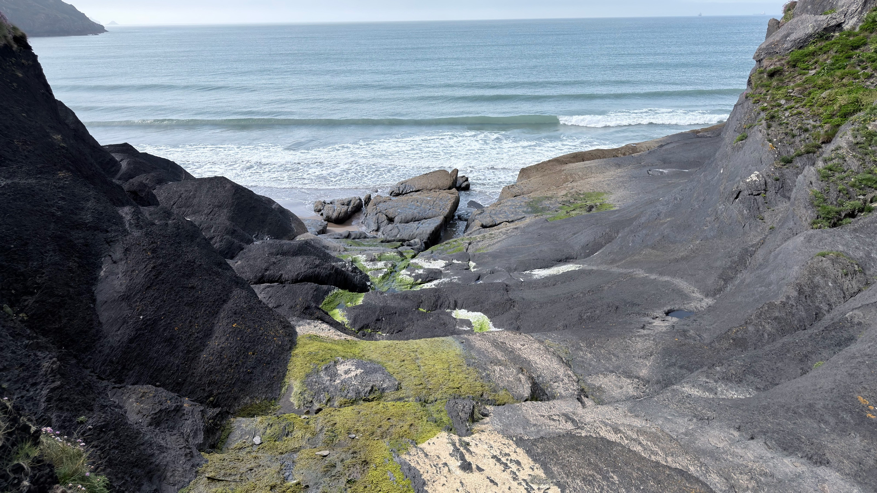 A rocky landscape leads down to a sandy beach and the ocean, with waves approaching the shore.