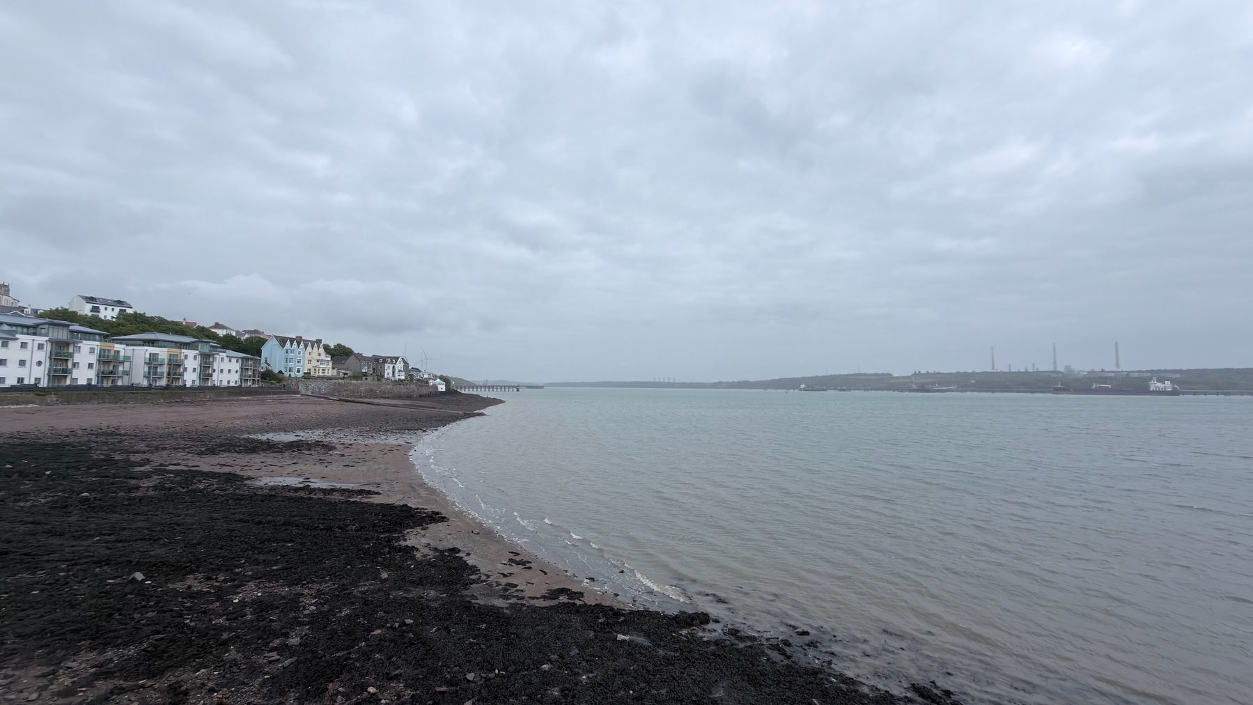 A cloudy, overcast day features a coastal scene with a shoreline, distant buildings, and the sea stretching into the horizon.