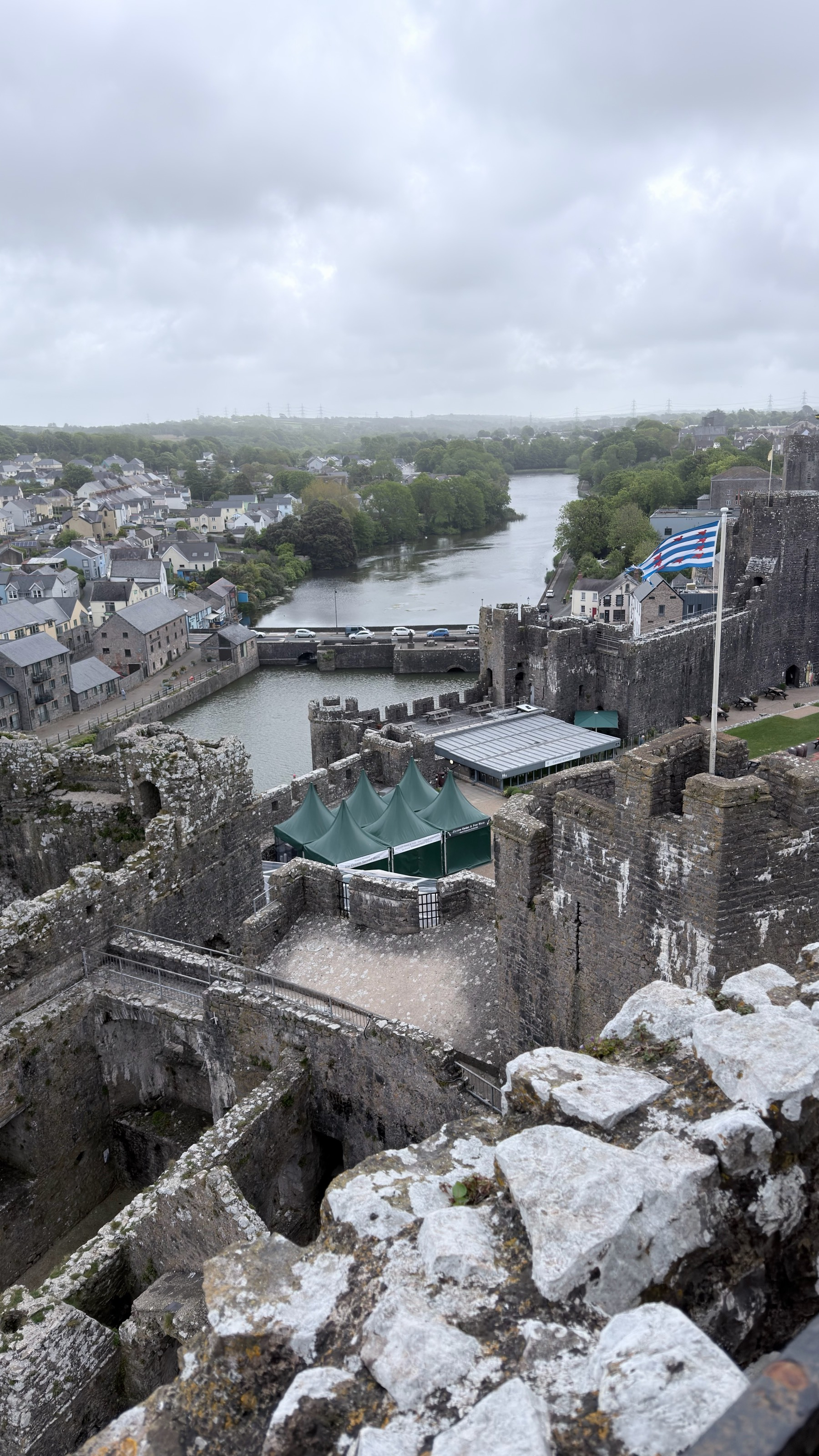A historic castle with stone walls and a river in the background under a cloudy sky.