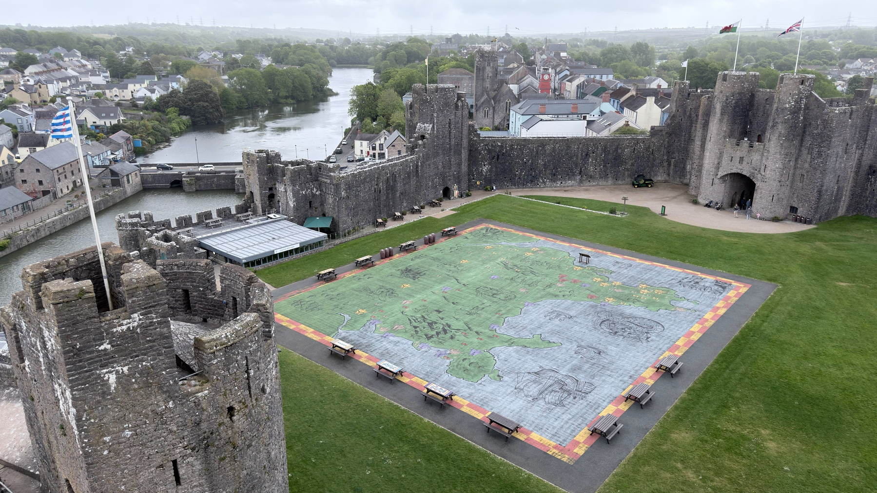 An aerial view captures a large, detailed map laid out on the grounds within the walls of a historic castle, surrounded by a river and a townscape.