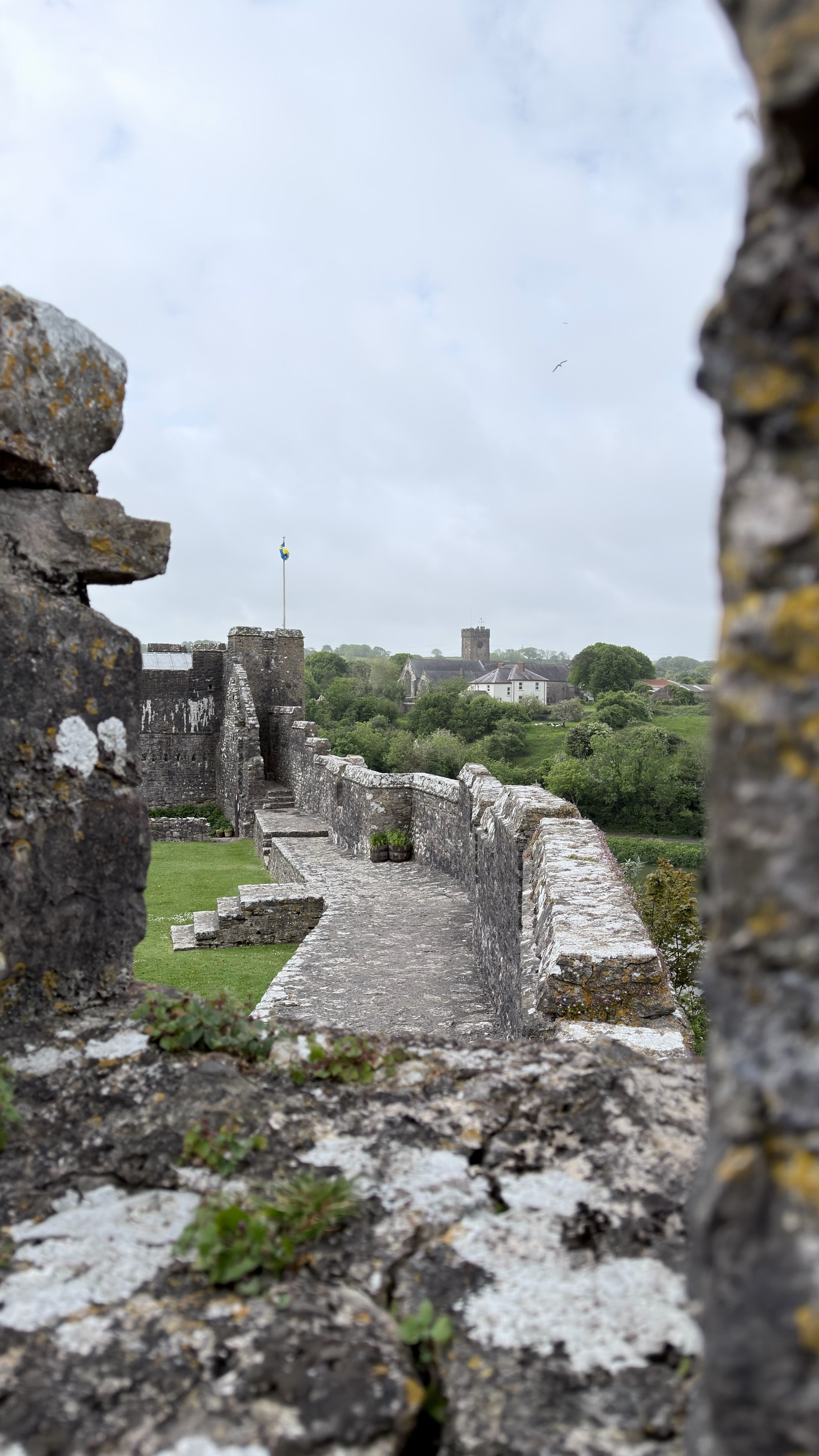 A view through a stone wall opening reveals a historic fortress with a pathway, greenery, and a church in the distance.