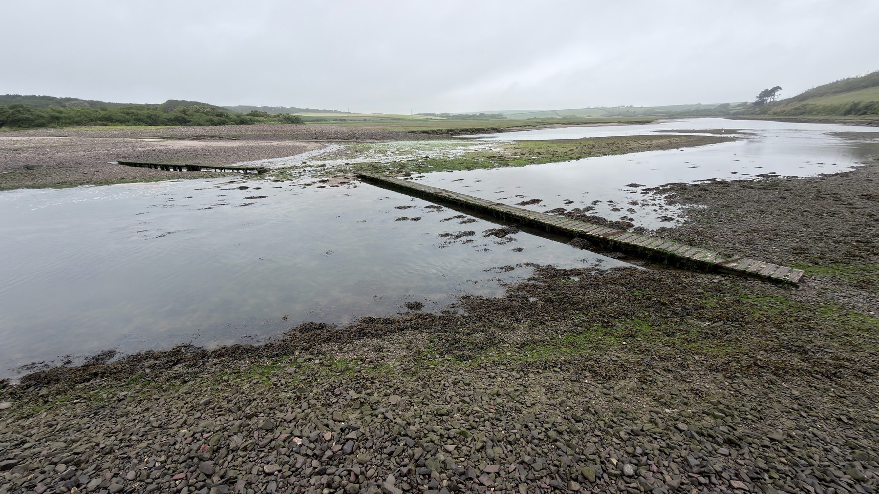 A rocky shoreline features a wide, shallow body of water with a narrow wooden walkway extending into it, set against a backdrop of overcast skies and distant greenery.