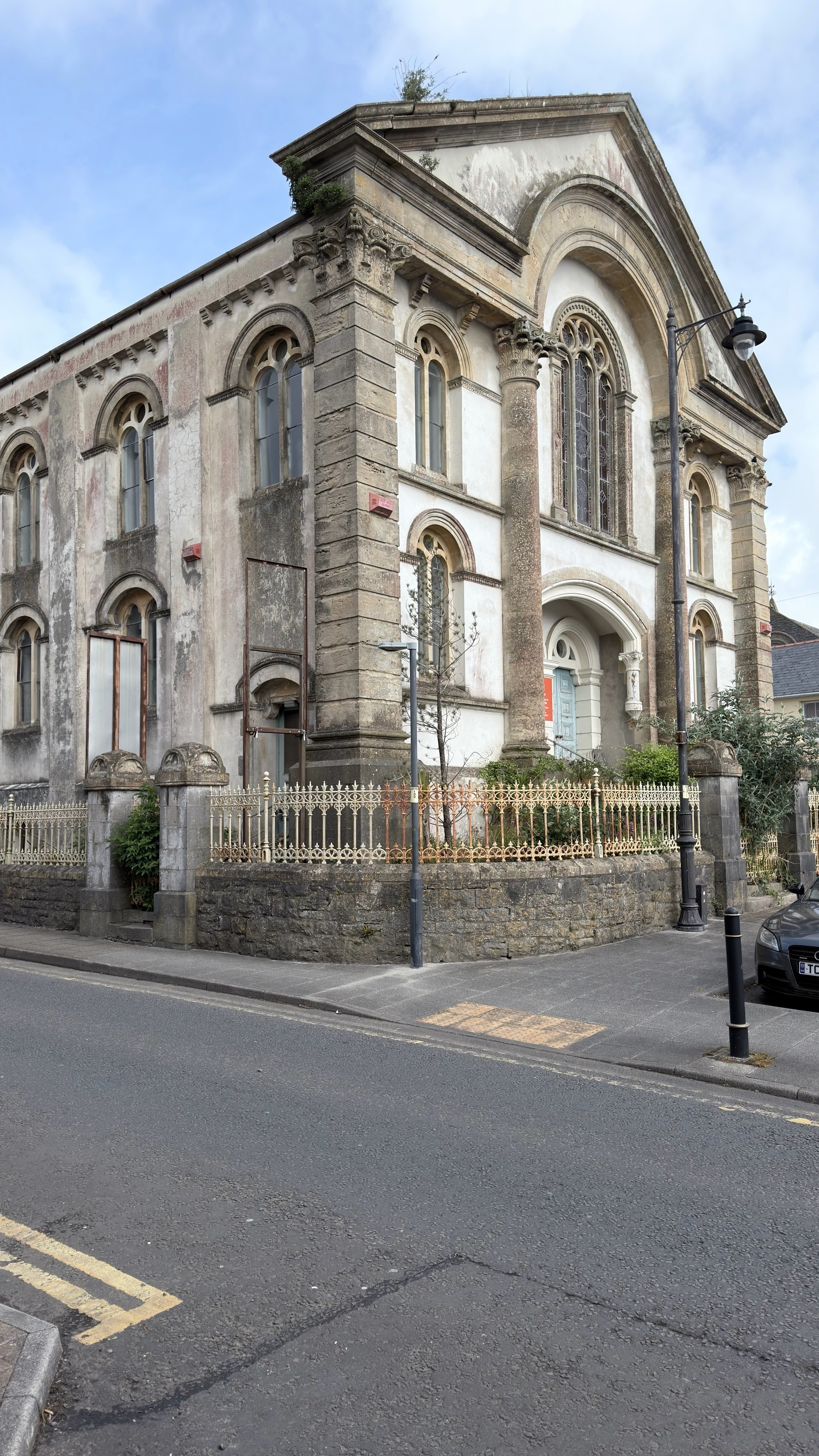 A historic, weathered building with arched windows and a fenced perimeter stands beside a street lined with yellow road markings.