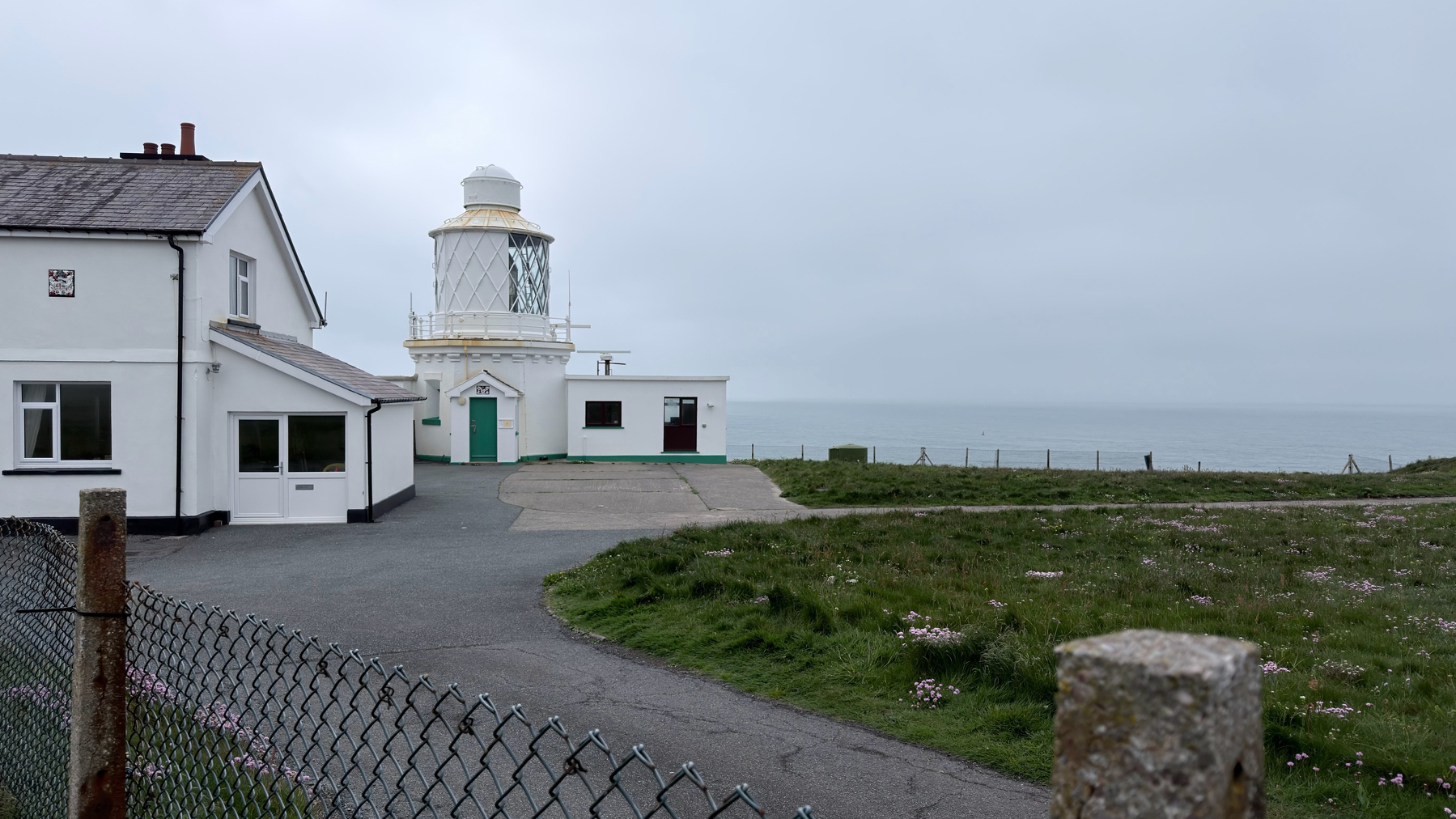 A white lighthouse with an adjoining building sits near the coastline under an overcast sky, surrounded by grassy land and a chain-link fence.