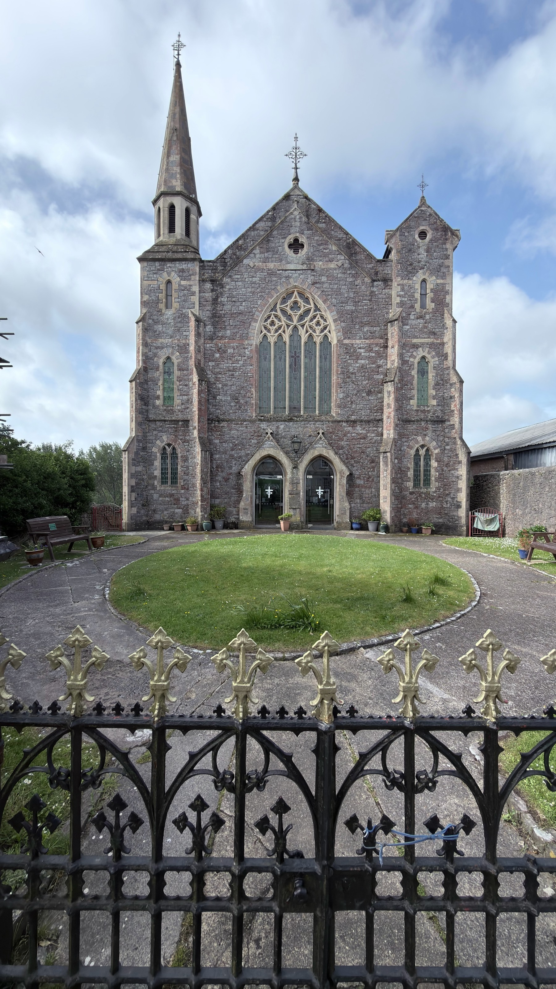 A historic stone church with arched windows and a tall spire stands behind an ornate wrought-iron fence, surrounded by a grassy area and benches.
