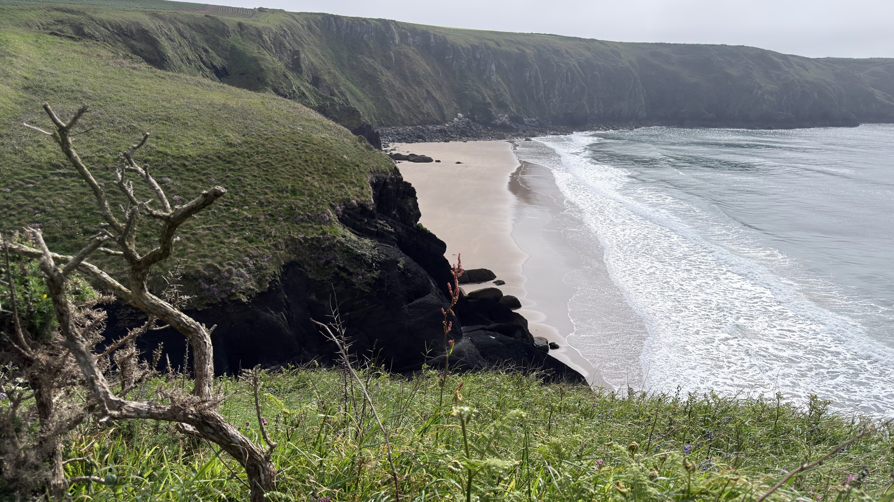 A scenic coastline features a sandy beach nestled between rocky cliffs and grassy hills.