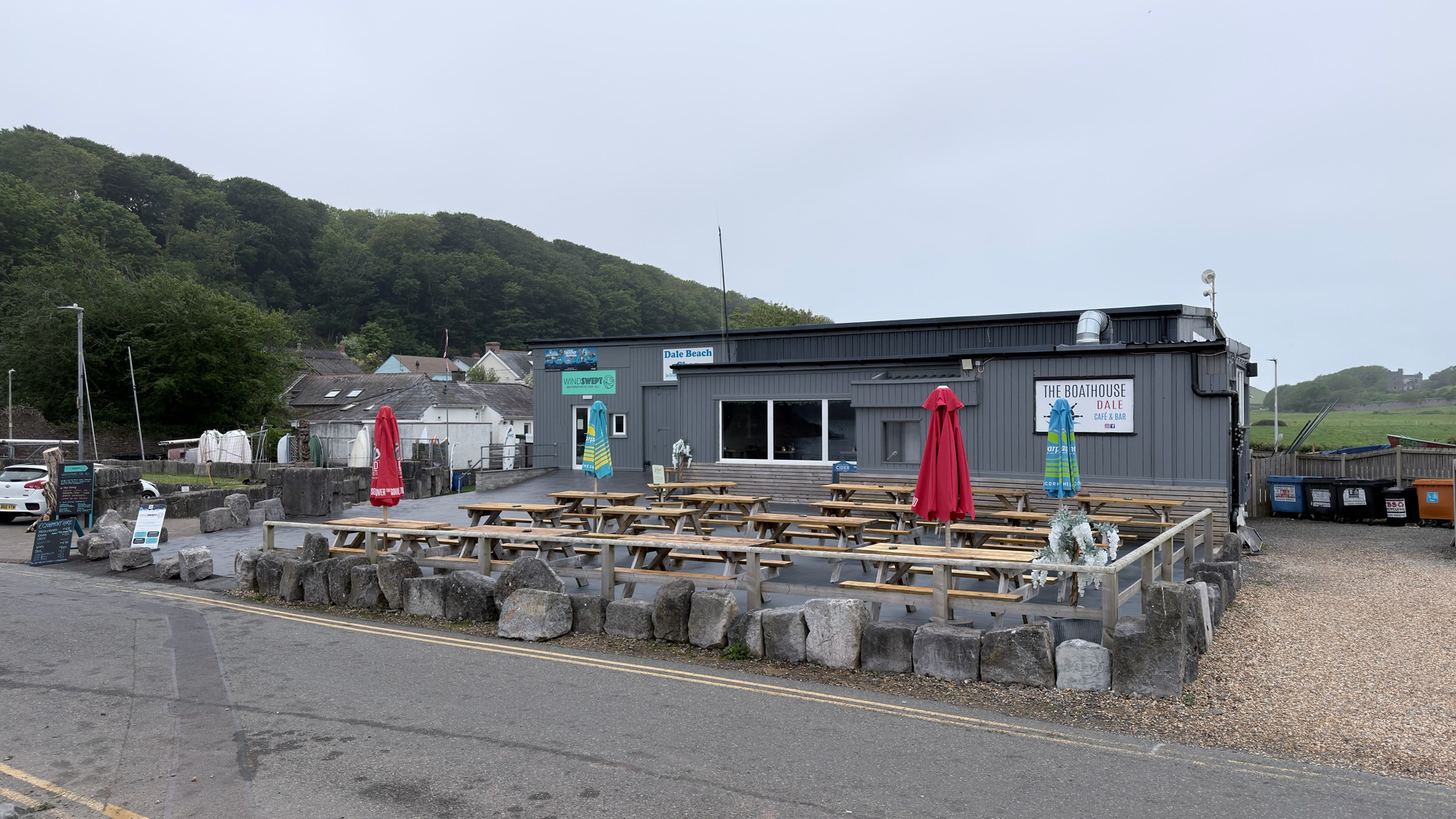 A seaside cafe with outdoor wooden picnic tables and colorful umbrellas is surrounded by a stone barrier, set against a backdrop of trees and nearby houses.