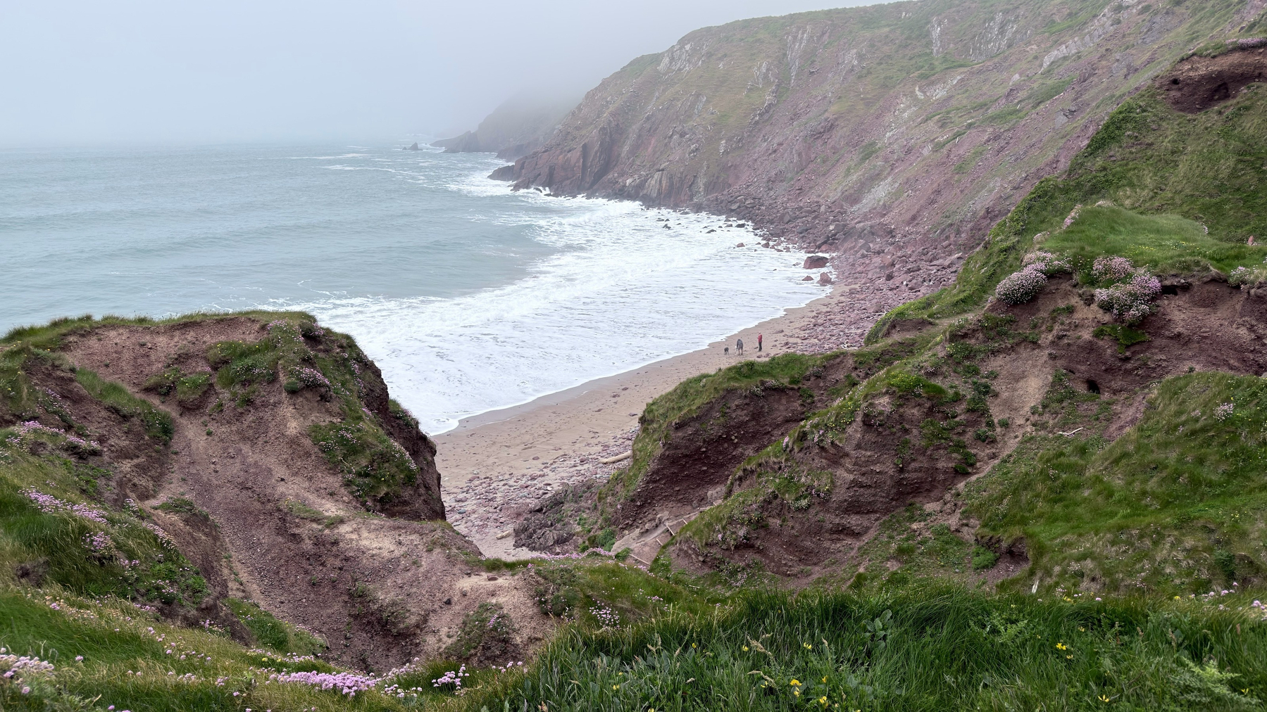 A misty coastal scene features rocky cliffs, a sandy beach, and waves gently crashing against the shore.