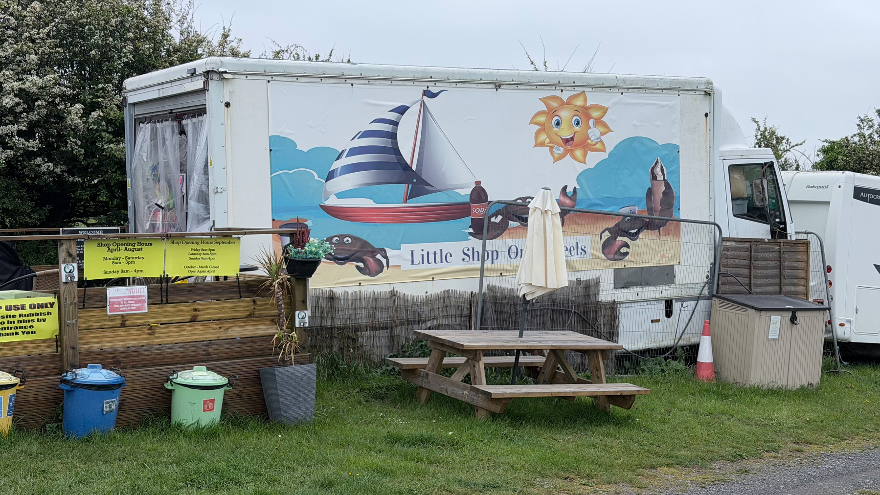A food truck named Little Shop of Shells is adorned with a nautical-themed mural, featuring a sailboat and smiling sun, and is surrounded by bins and picnic tables.
