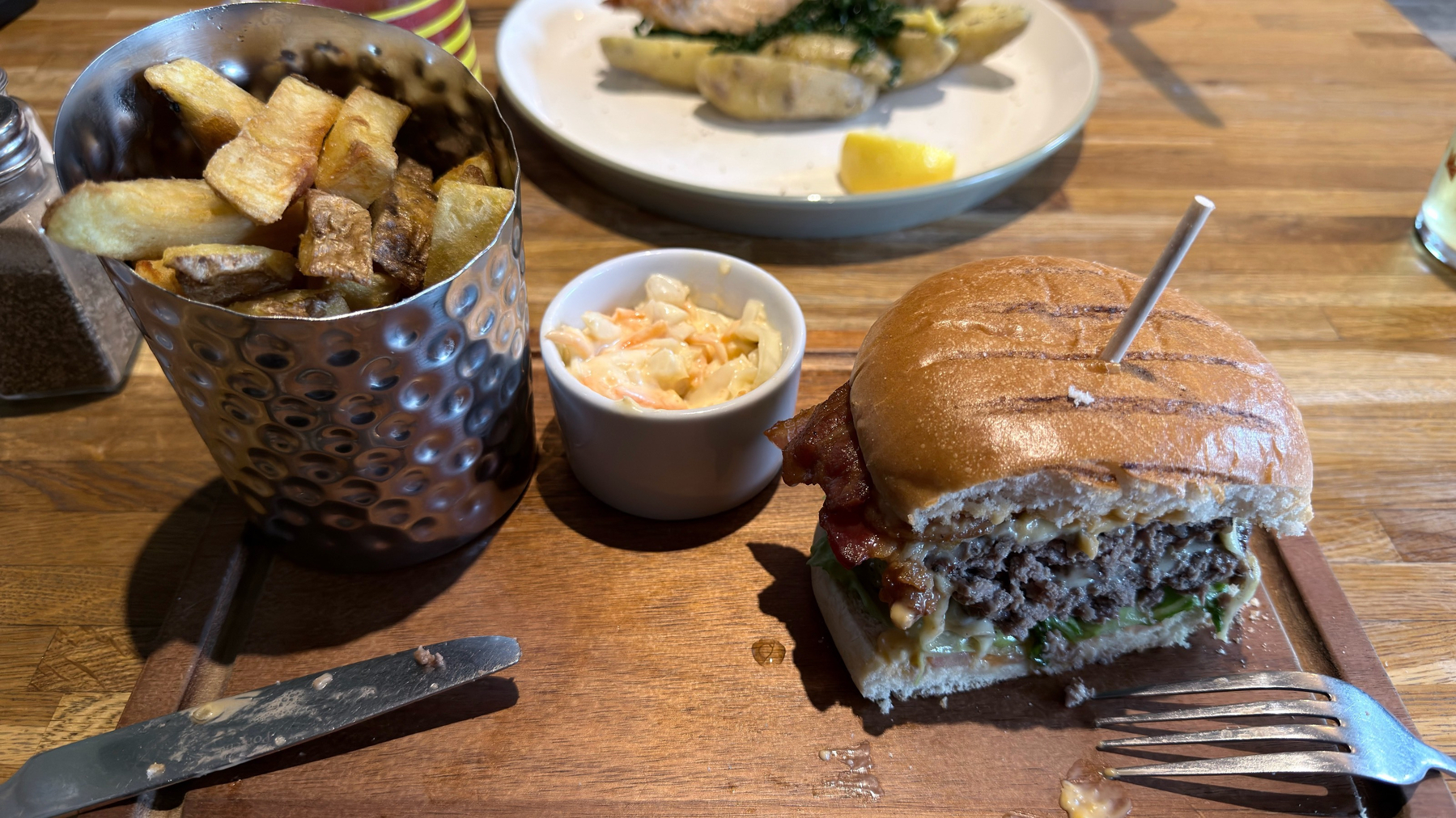 A hamburger with a toothpick, a cup of coleslaw, and a metal bucket of fries are presented on a wooden board next to cutlery, with another dish blurred in the background.