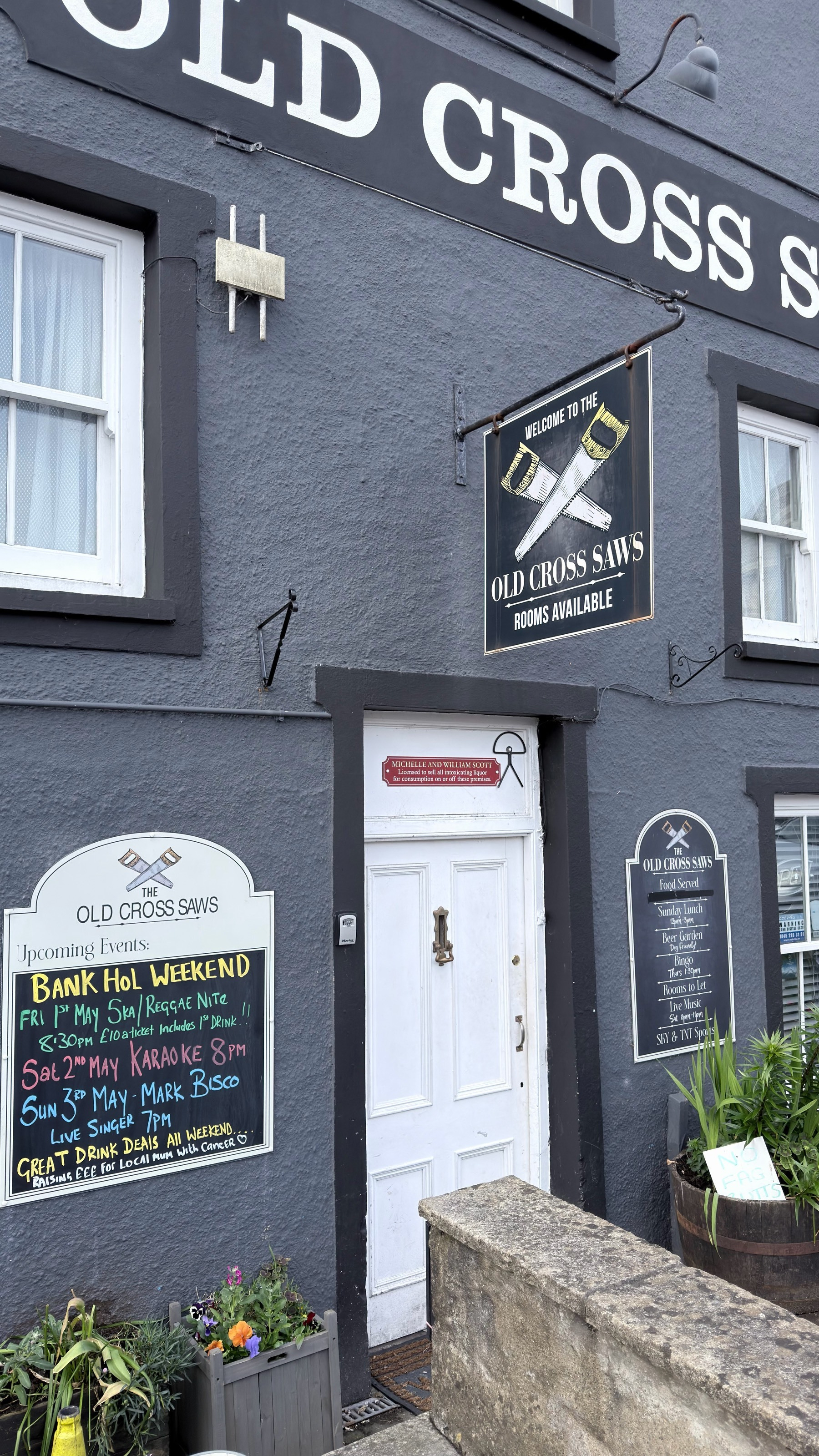 A dark gray building with signs for Old Cross Saws, displaying event information and a welcoming entrance with flower pots nearby.