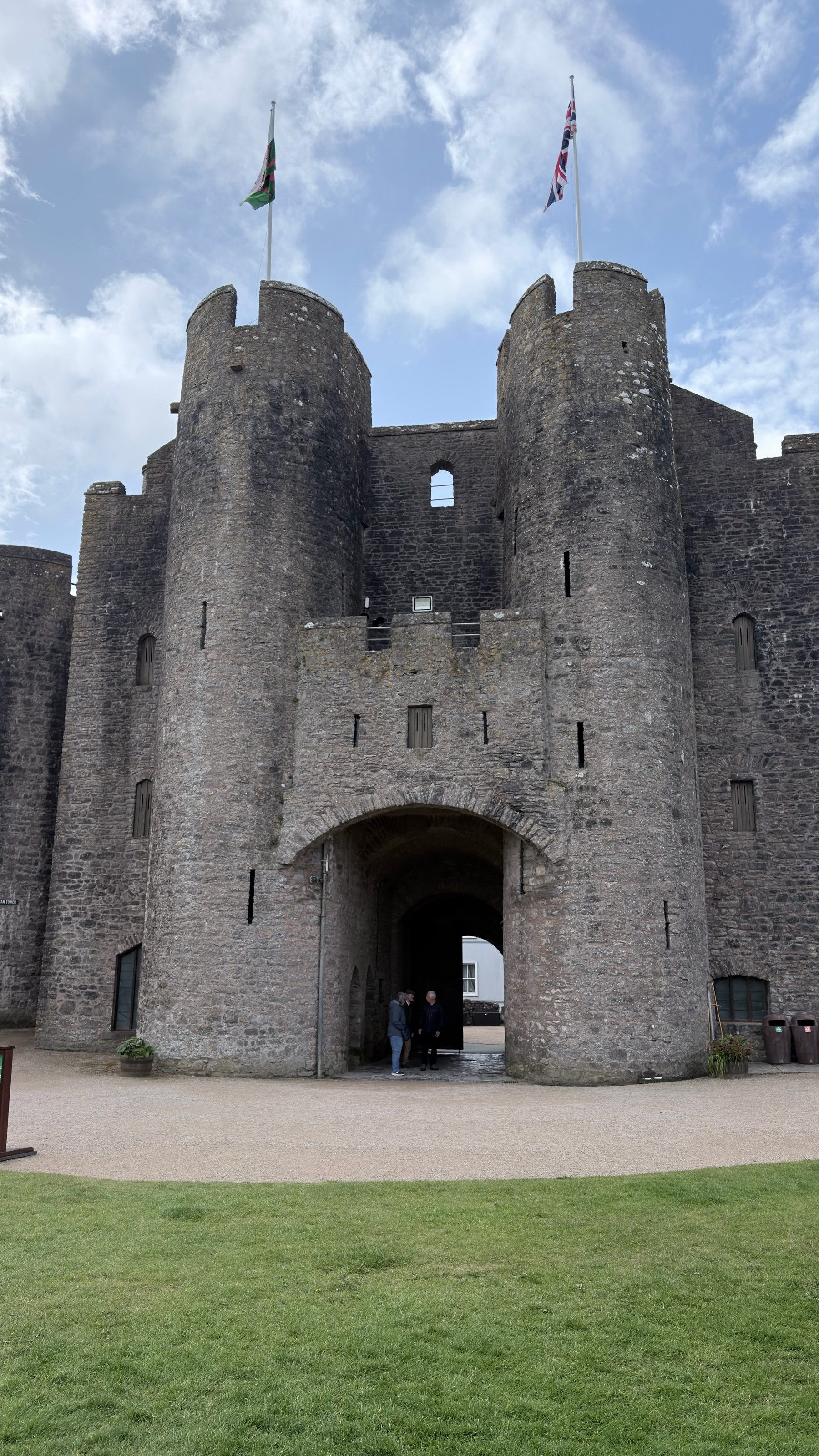 A large stone castle with two flags flying on top stands under a partly cloudy sky, with a pathway leading to its entrance.