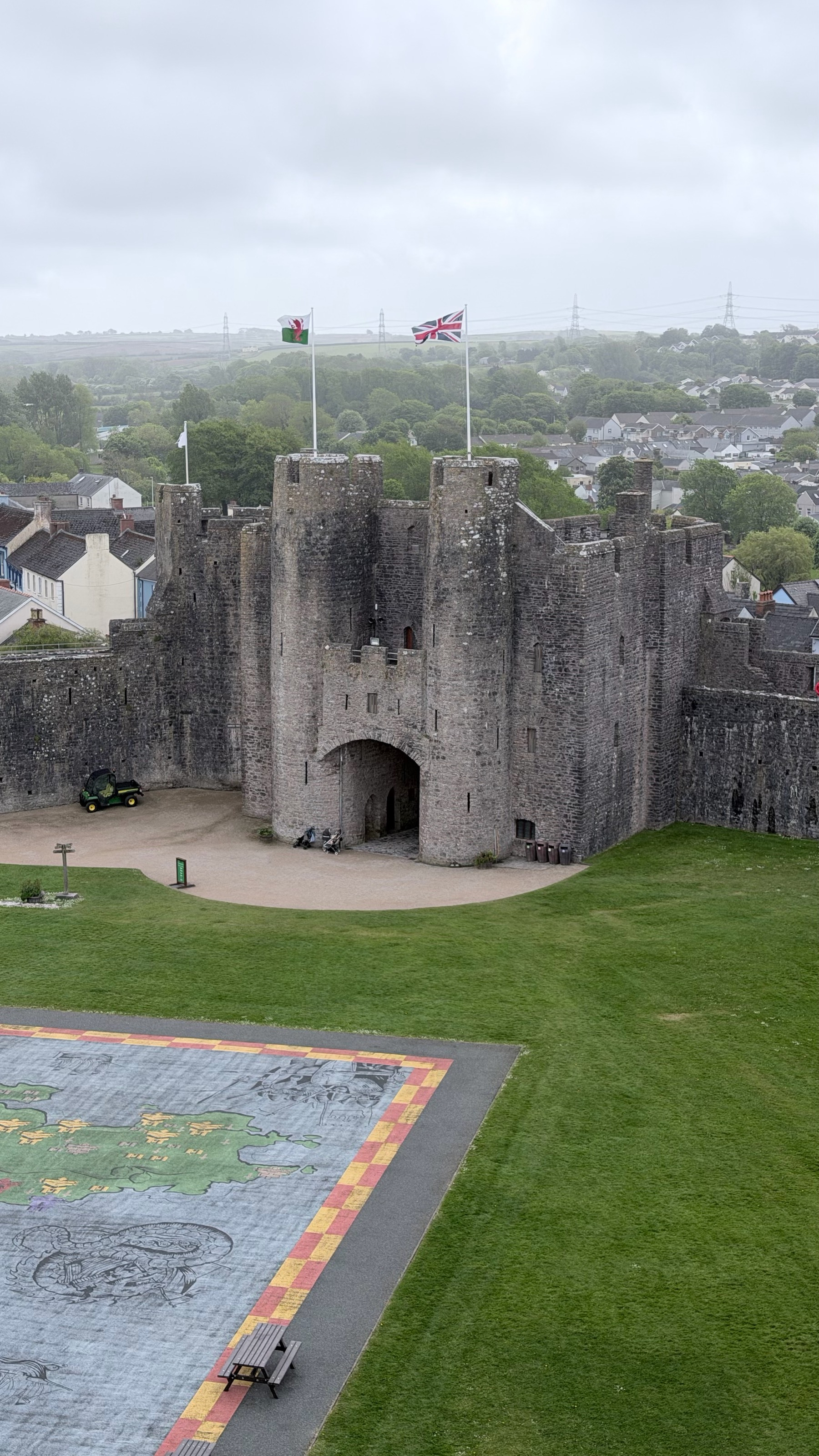 A historic stone castle with two flags flying above is surrounded by a green lawn and mural near a residential area under a cloudy sky.